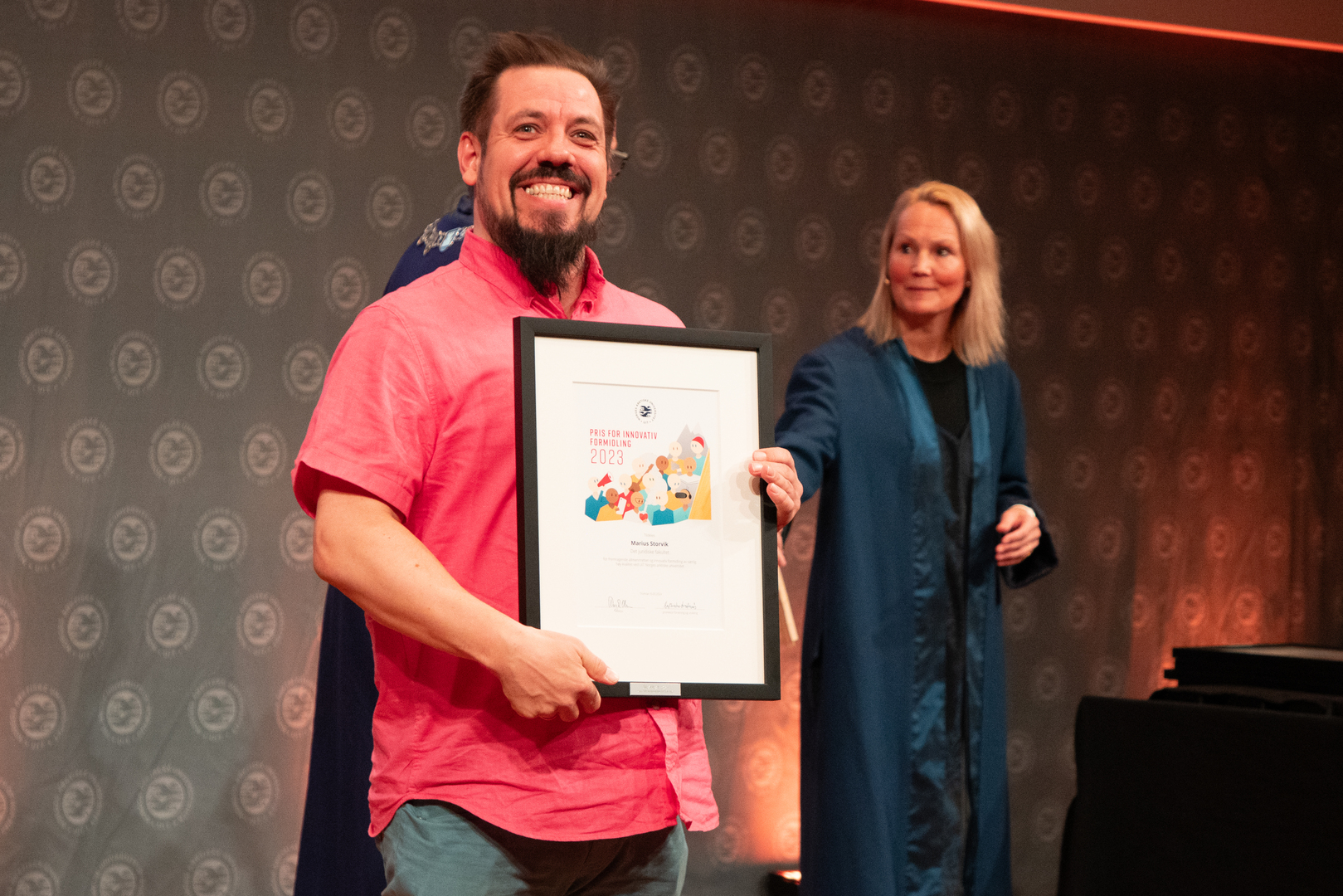 arsfest_2024_UIT_24200_Foto_Jorn_Berger_Nyvoll.jpg Mann i rosa skjorte mottar diplom, kvinne i blå kappe står i bakgrunnen.