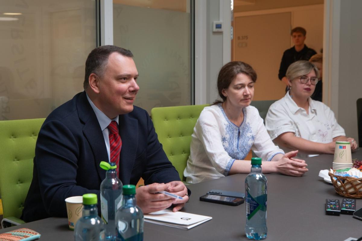 a man talking at a meeting. two women are sat next to him, listening