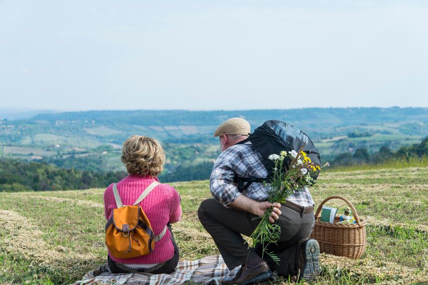 Et eldre par sitter p&aring; et teppe p&aring; en slette med ryggen til og ser ut over et gr&oslash;nt landskap. Mannen har en bukett med blomster bak ryggen.