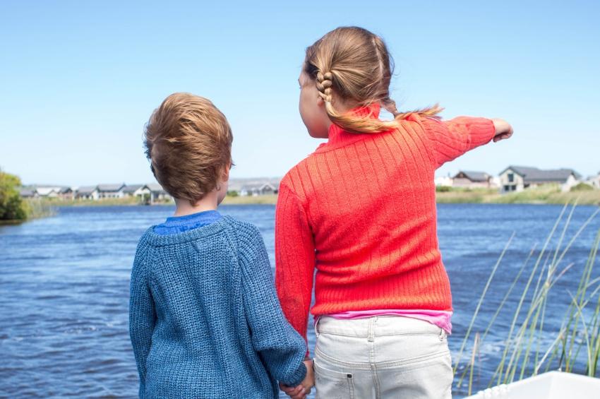 To barn st&aring;r p&aring; en brygge og holder hender. Den eldste som er en jente peker utover vannet. Hun er kledd i r&oslash;dt. Det yngste barnet er kledd i bl&aring;tt.