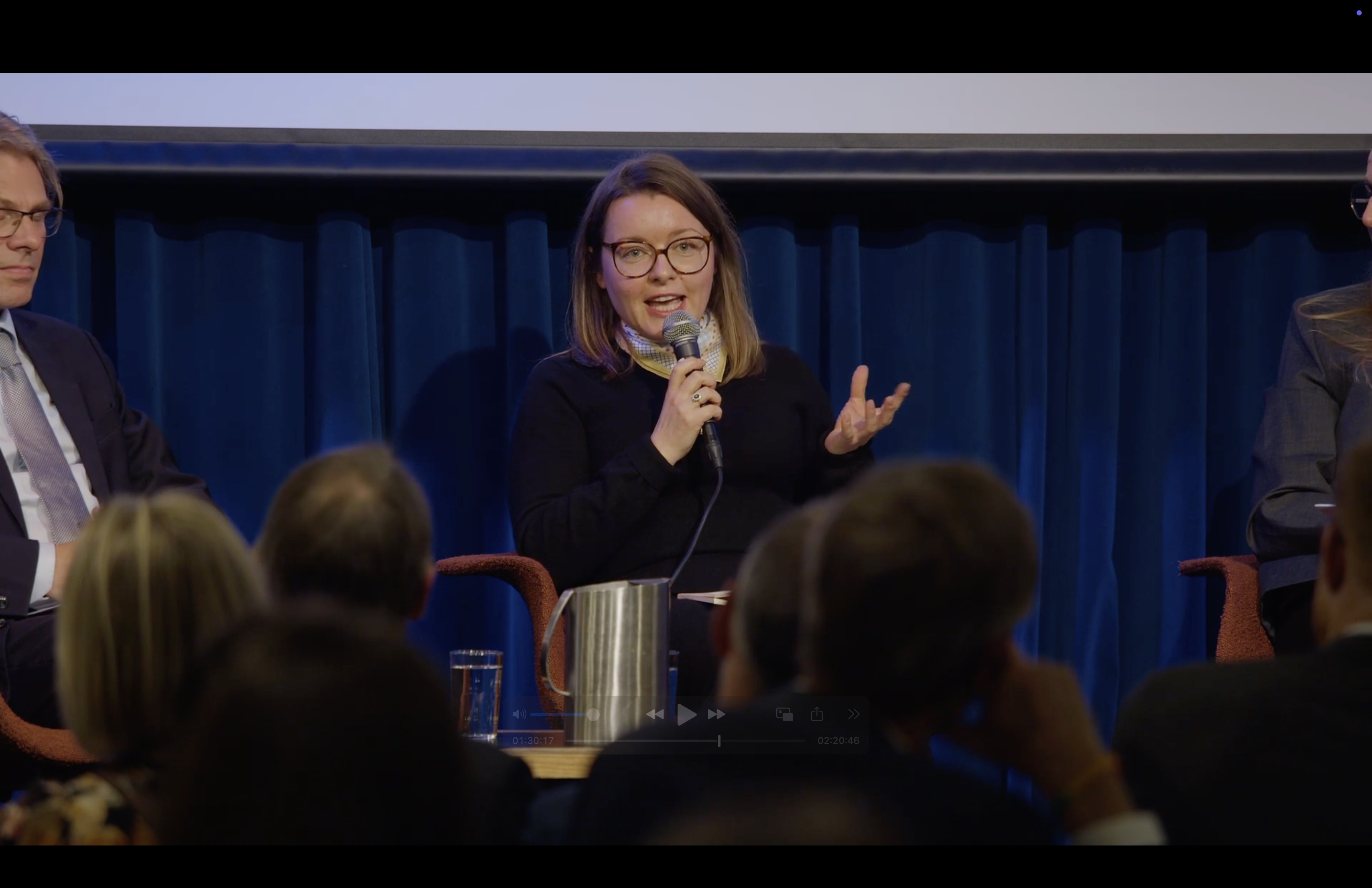 woman speaking into a microphone, sitting in a chair on a stage. 