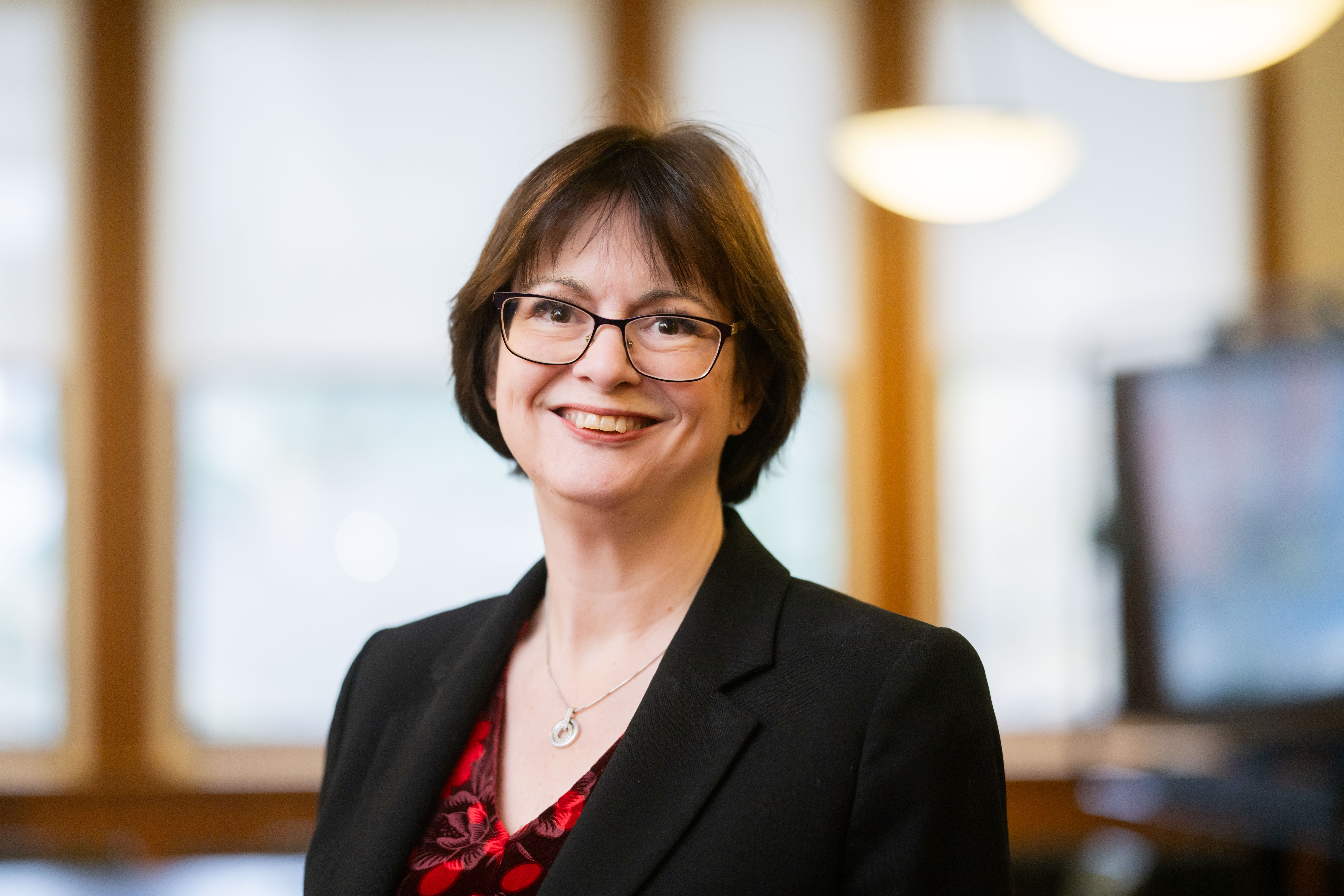 woman, short brown hair, looking into the camera, portrait photo, smiling, wearing red top with flowers and black blazer, glasses on, silver necklace