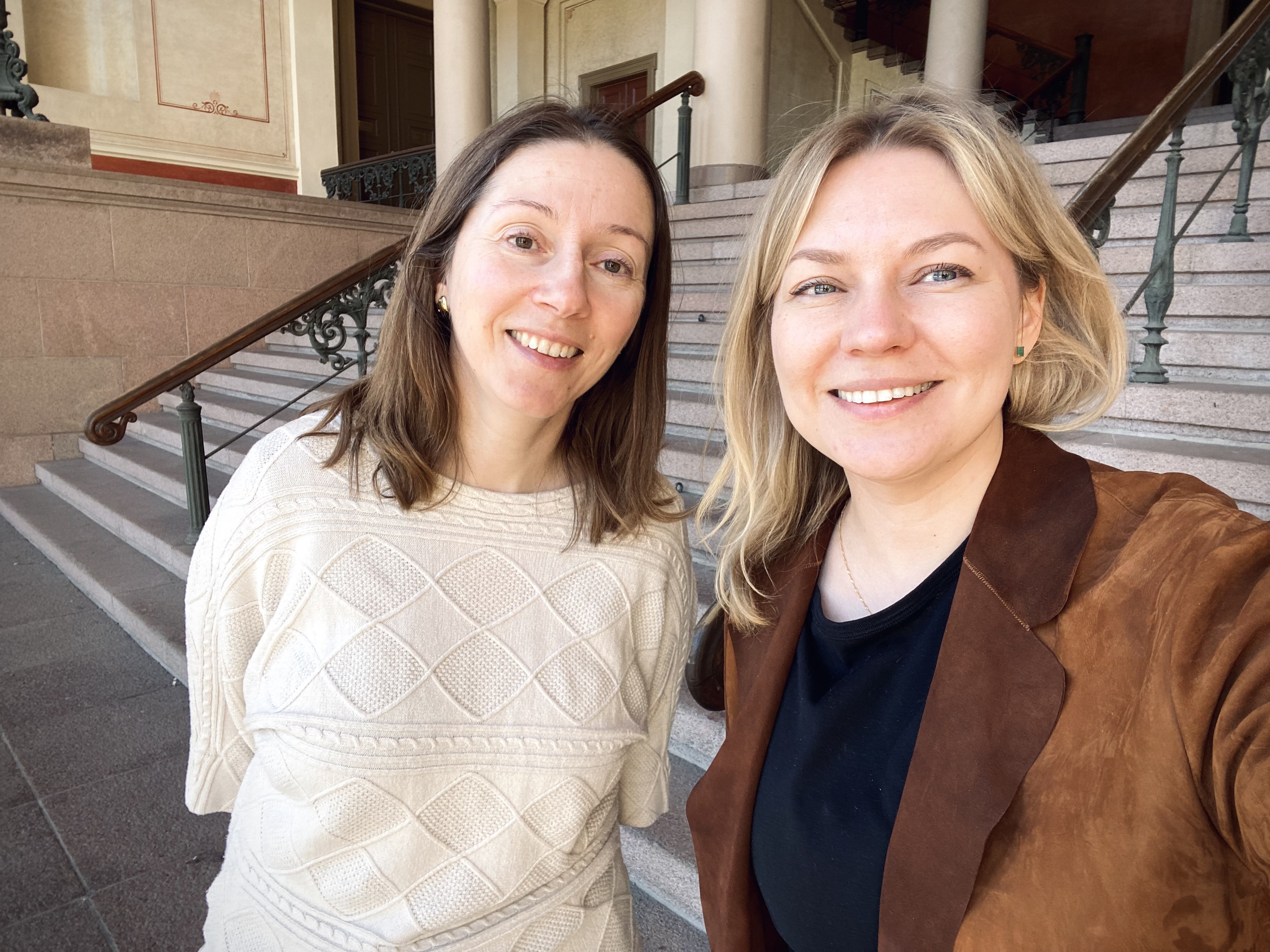 two women, looking inot the camera, one with brown hair and the other with light, blond hair, both are smiling