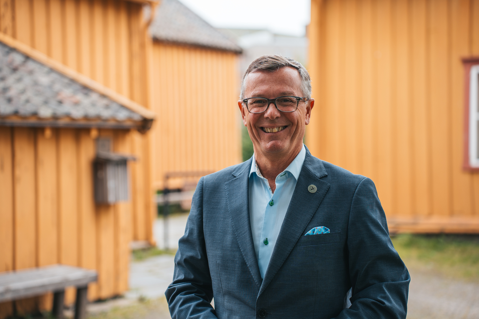 Rektor i dress i forgrunnen av eldre bebyggelse i oransje farge. 